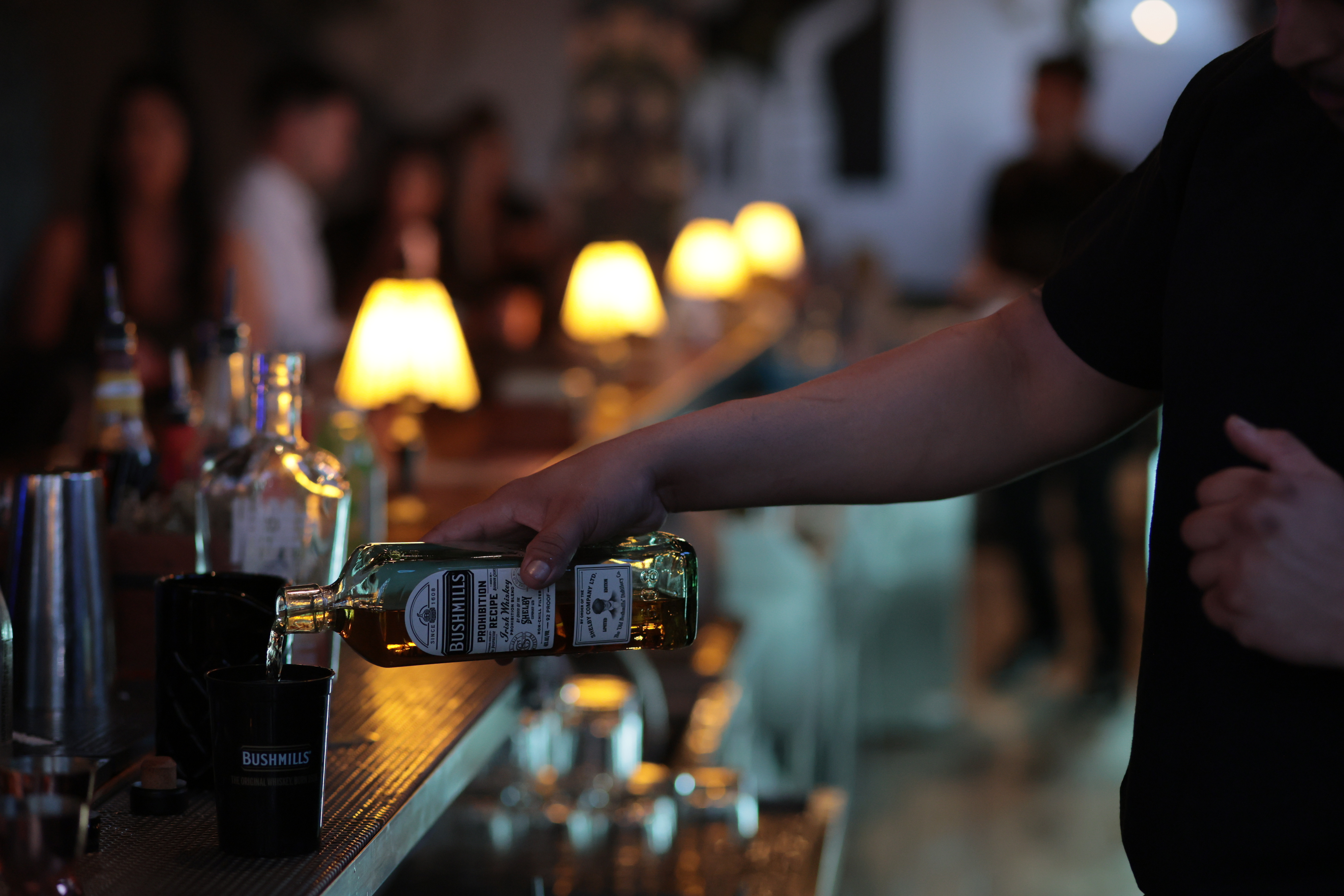 A bartender pouring a bottle of Bushmills whiskey at a bar, with a second bartender, guests, and a row of lights receding down the bar